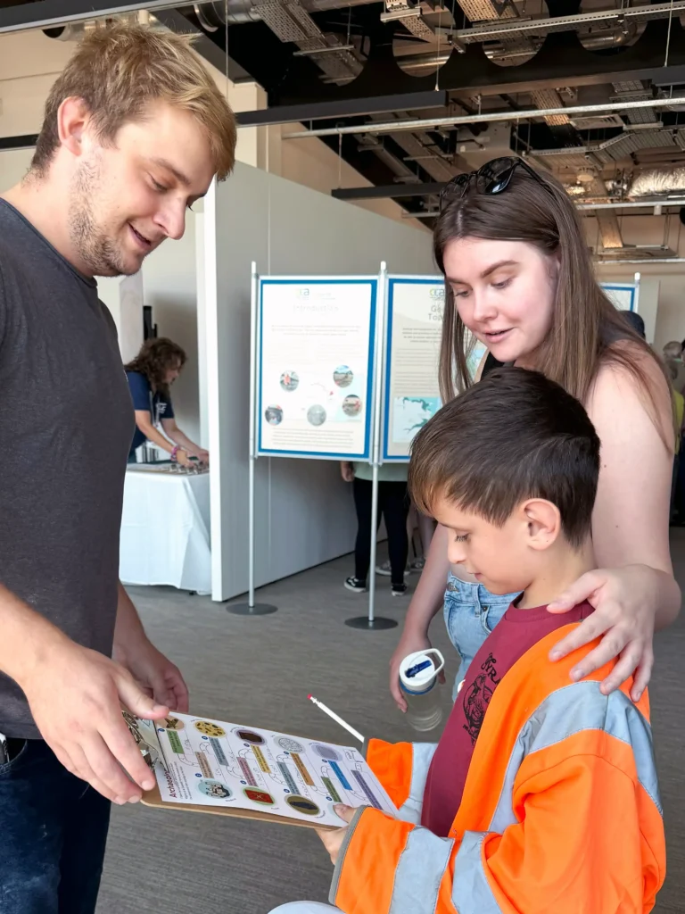 A man and woman lean over a clipboard held by a young boy wearing a bright orange safety vest, looking at the sticker trail