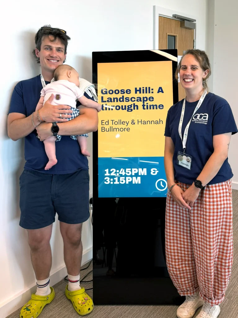 A man holds a baby next to a digital display that reads "Goose Hill: A Landscape through time" with scheduled times of 12:45 PM and 3:15 PM, and a woman stands to the right of the display.