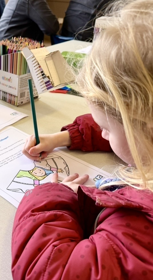 A young child with blonde hair, wearing a red jacket, is concentrating as they color in a picture with a green pencil.