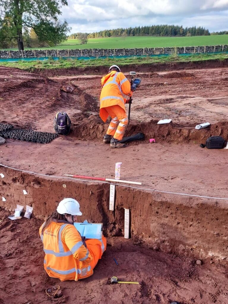 Archaeologists wearing orange highvis PPE. One is sat on the ground holding a board with blue paper recording. The other is stood with GPS.