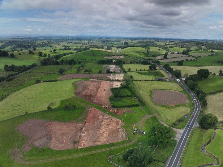 An aerial view of the A66 excavations running next to the route of the A66 roadway. Both road and excavation are nestled in amongst a patchwork of green fields framed at the top by a slightly cloudy blue sky.